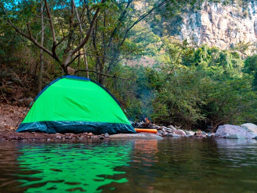 Green dome tent pitched next to a body of water surrounded by trees and cliffs in background