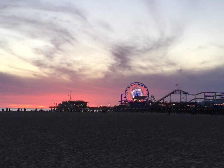 Dimly lit beach sand leading up to a silhouette of a ferris wheel and roller coaster on a pier with a pink sunset sky