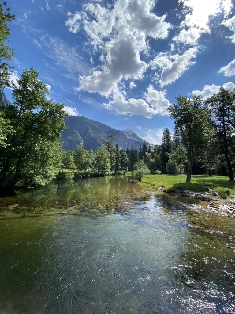 Calm Merced River surrounded by green grass and trees with mountains, blue sky, and scattered glowing clouds