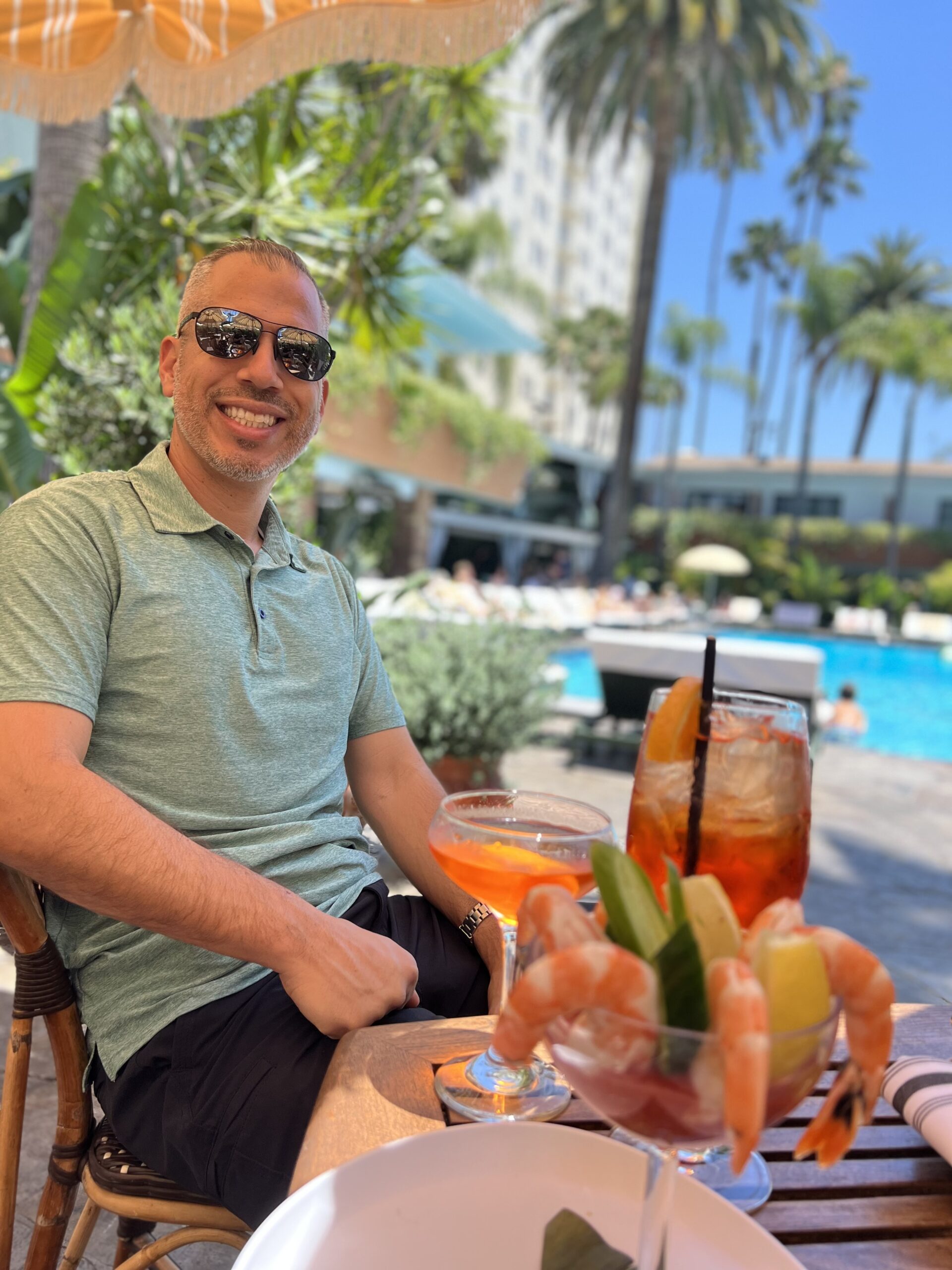 The author, Kevin, sitting and smiling at a table with drinks and a shrimp cocktail by the Hollywood Roosevelt Hotel pool