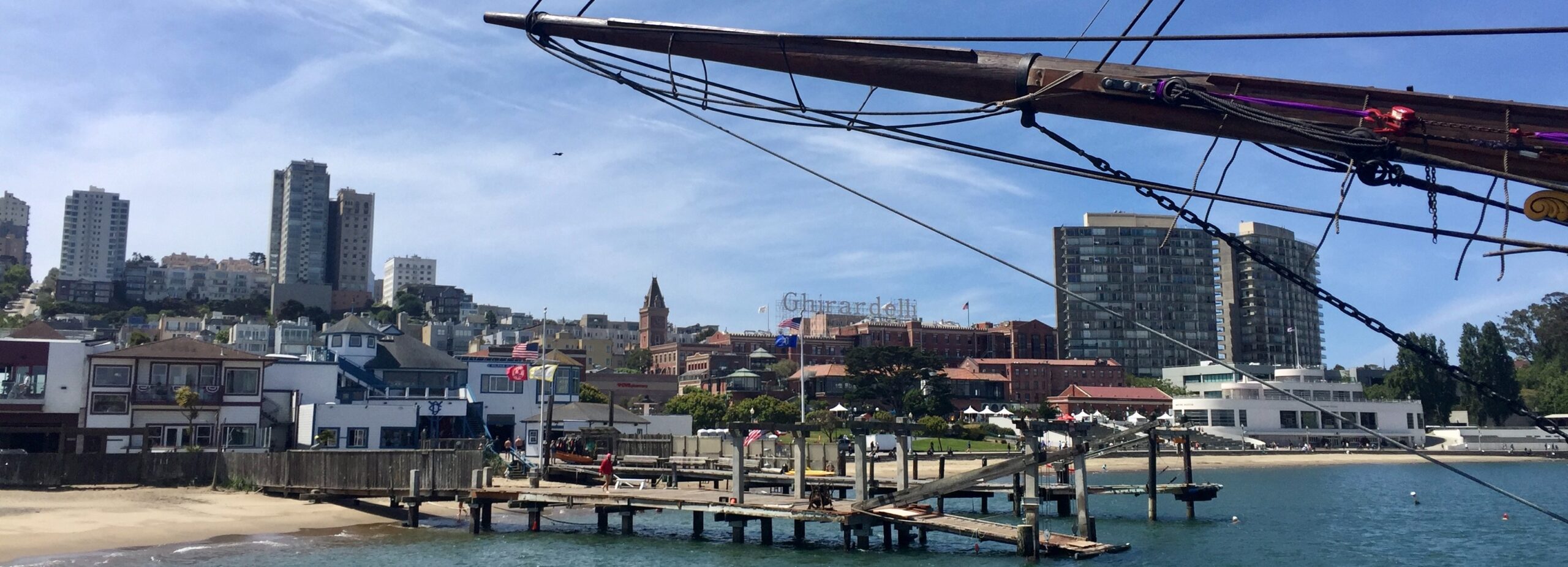 Sail boat mast over water with a small deck and several San Francisco buildings in background