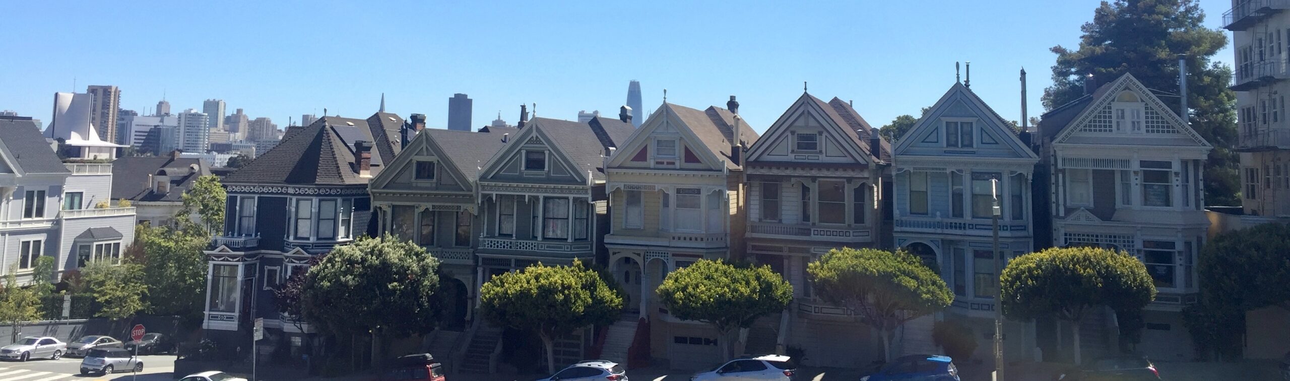 Seven different colored Victorian homes with green trees in front and San Francisco skyline and blue sky behind