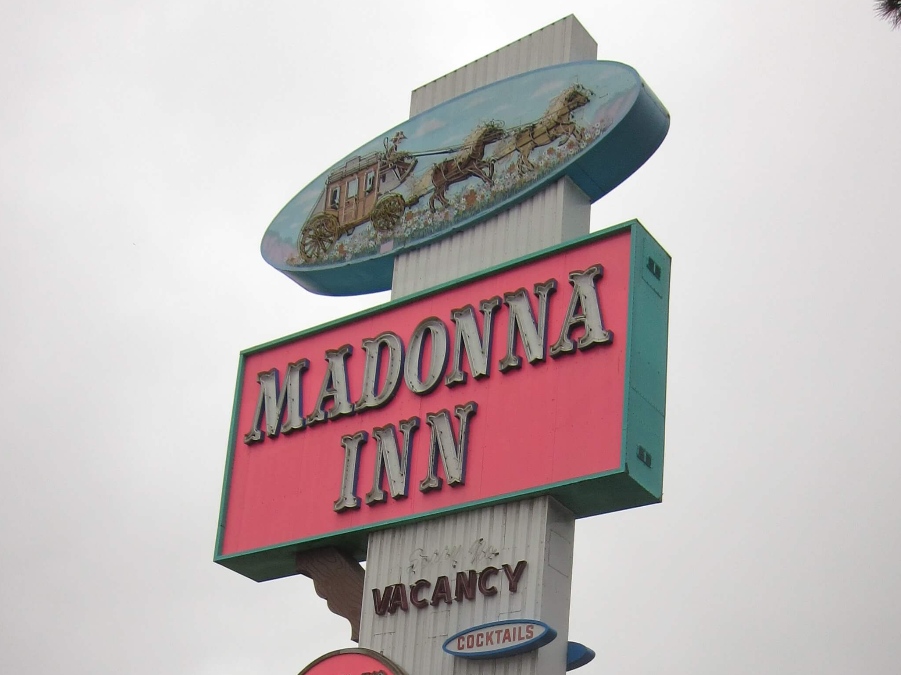 Pink and white Madonna Inn sign with a horse carriage artistic unlit light at the top. Link to Where to Stay on page