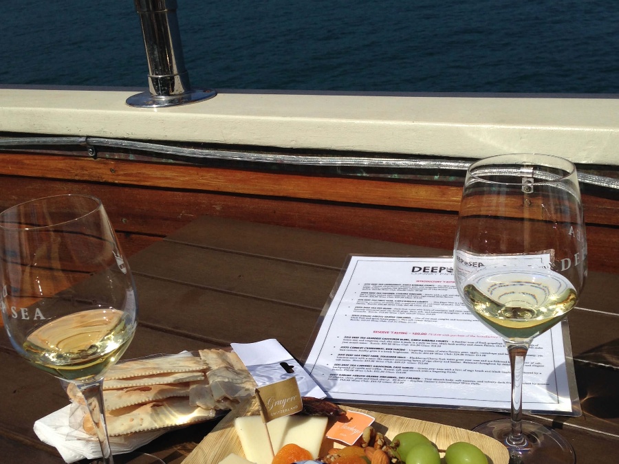 Two glasses of white wine on a table with cheese, fruits, crackers, and a menu with the ocean in the background