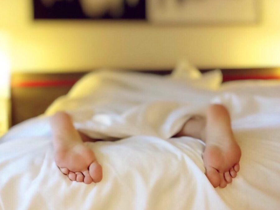 Two bare feet sticking out of white sheets on a bed of a person laying down on their stomach in a hotel room