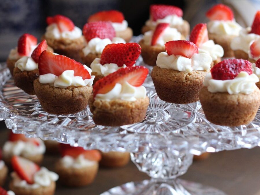 Crystal platter holding several strawberry iced mini cakes with more mini cakes in background