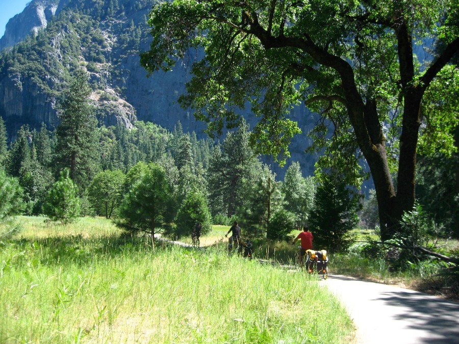 Back of two bicyclists riding a paved trail through green grass surrounded by trees and rocky mountains in the background