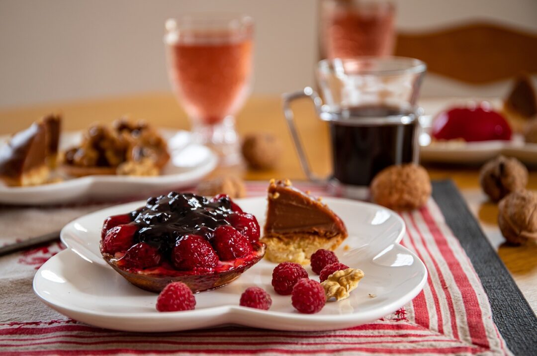 Raspberry fruit tart on a plate with more raspberries and cake slice with drinks and more treats blurred in the background