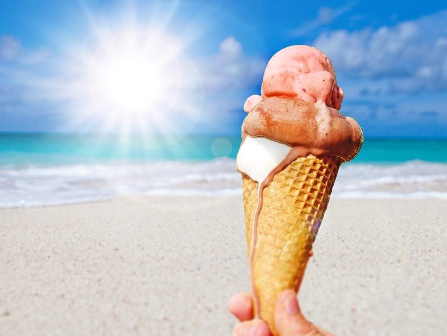 A hand holding a two scoop ice cream cone dripping with blurred sand, beach, and sky in background