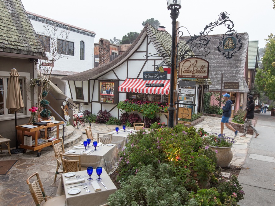 Outdoor dining tables with blue glasses next to green bushes facing the street surrounded by folksy architecture