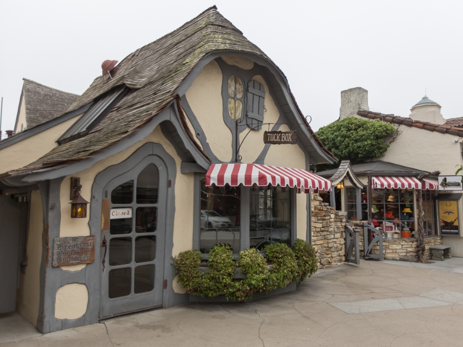 Fairytale style beige cottage with curvy wooden roofing and door and red and white window awning and green bush below window