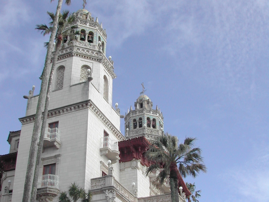 Side upward view of Hearst Castle front towers with palm trees in foreground and blue sky. Link to Hearst Castle page.