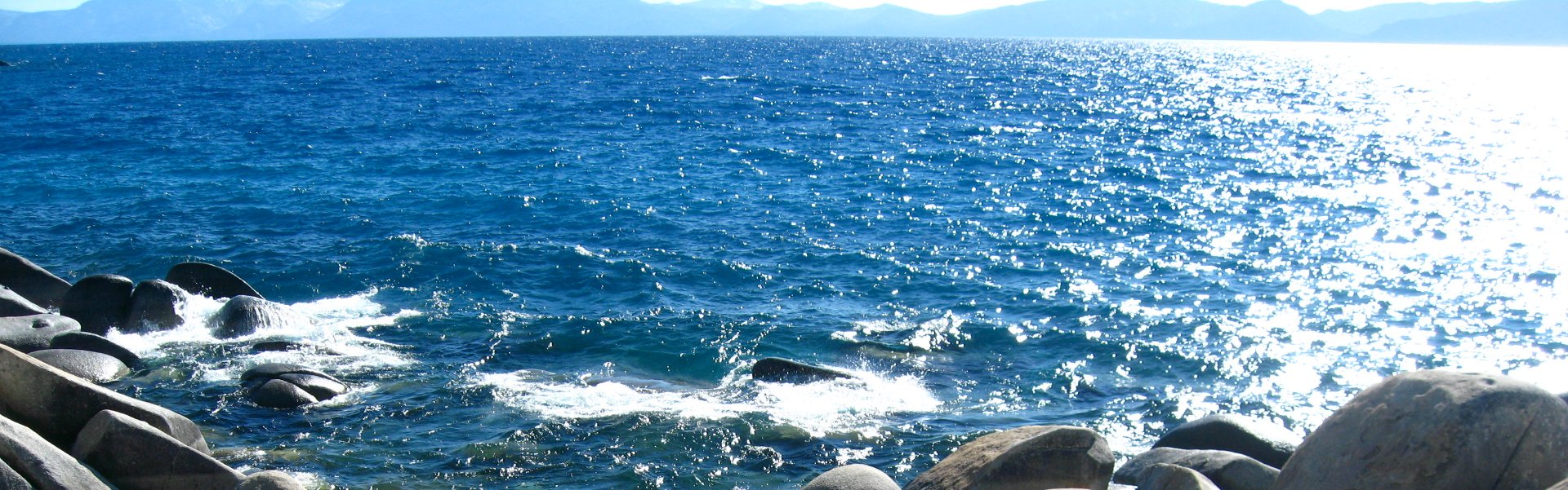 Smooth large boulders on the shore of deep blue Lake Tahoe with mountain range in background.