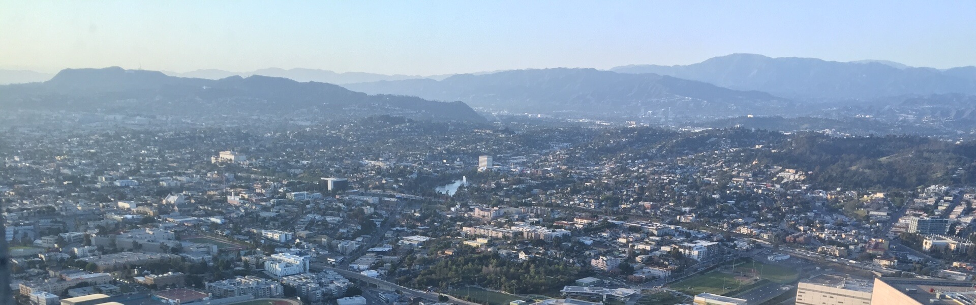 Panoramic view of Los Angeles from a downtown skyscraper building. Several buildings, trees, and hills in view.