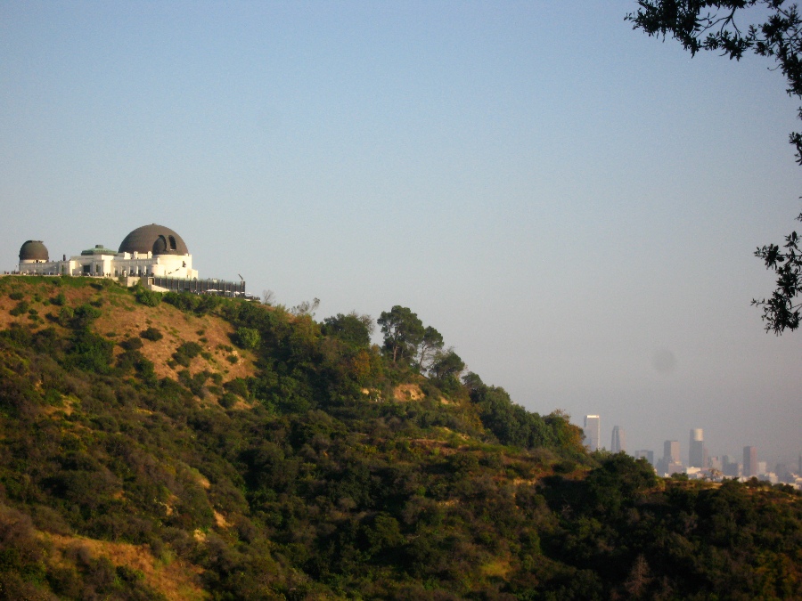 Exterior of Griffith Observatory sitting atop a green and brown hill overlooking downtown Los Angeles skyline