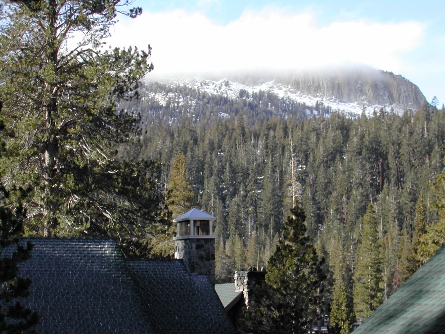 Cabin tops and tall green trees in front of a snow covered mountain on a cloudy day. Link to Mammoth Lakes page.