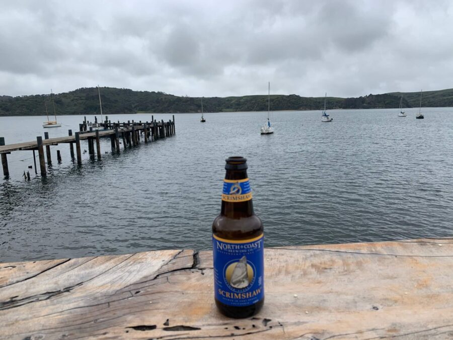 Opened Scrimshaw beer bottle on wooden counter overlooking Tomales Bay with a dock and boats.