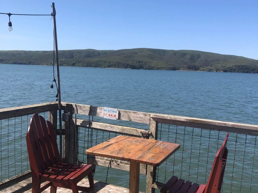 Rustic table and chairs outside on wooden deck overlooking Tomales Bay and green hill. Link to Where to Eat on page.