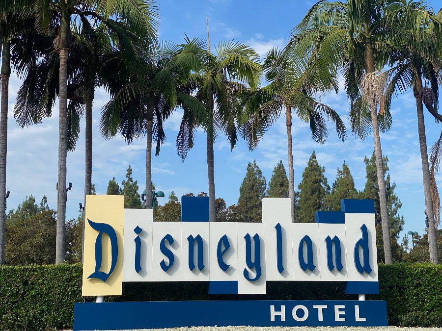 The Disneyland Hotel sign in white and blue with palm trees and blue sky in the background. Link to Where To Stay on page.