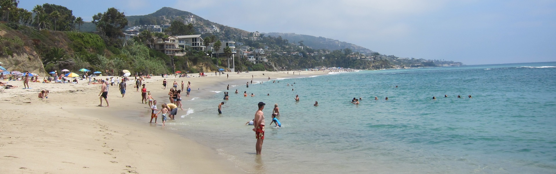 Soft sandy beach with people playing in the light blue water. Green hills and homes overlook the beach.