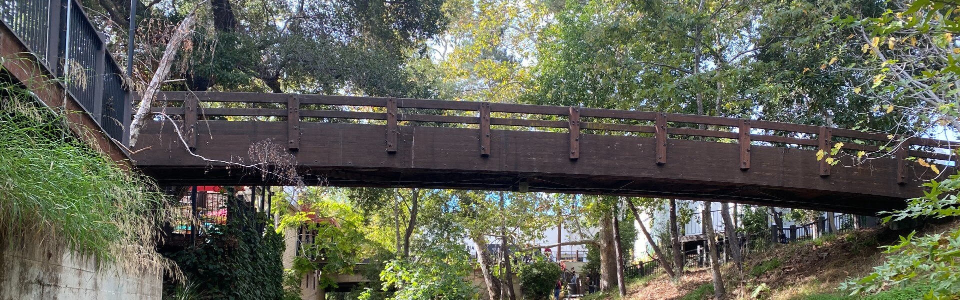 Brown wooden bridge over San Luis Obispo Creek surrounded by green trees.