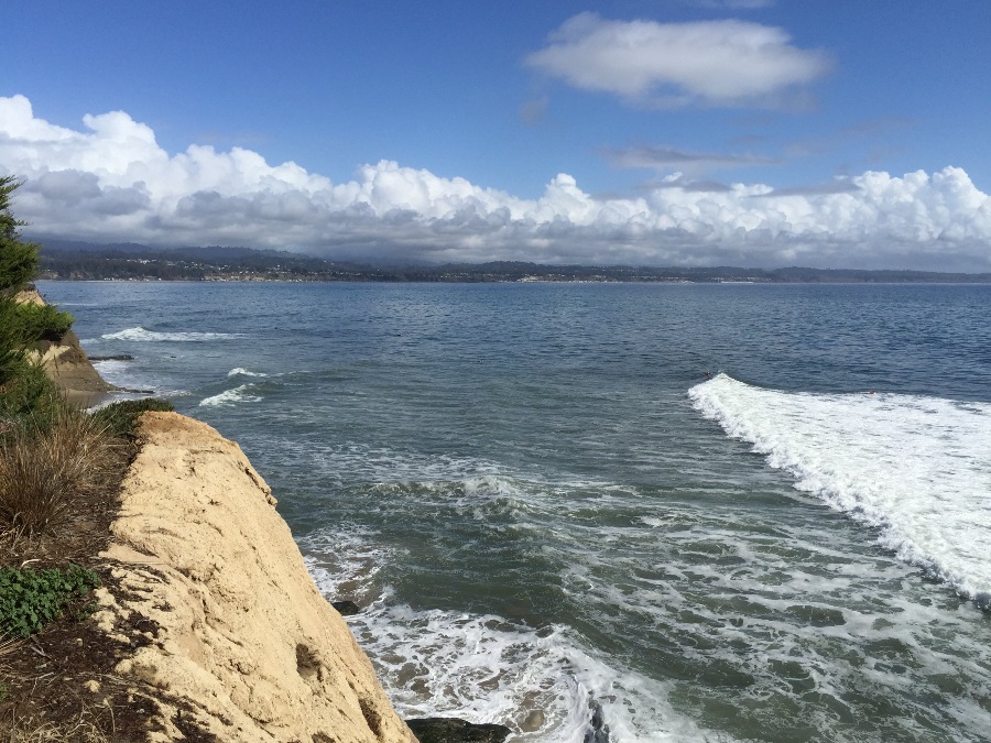 Ocean waves rolling towards Santa Cruz shore with hills, thick fluffy clouds, and blue sky in background.