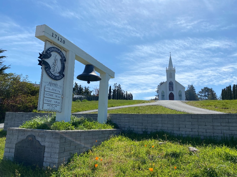 Wooden sign and bell in front of a white gothic chapel and blue sky with faint clouds. Link to Sonoma County page.