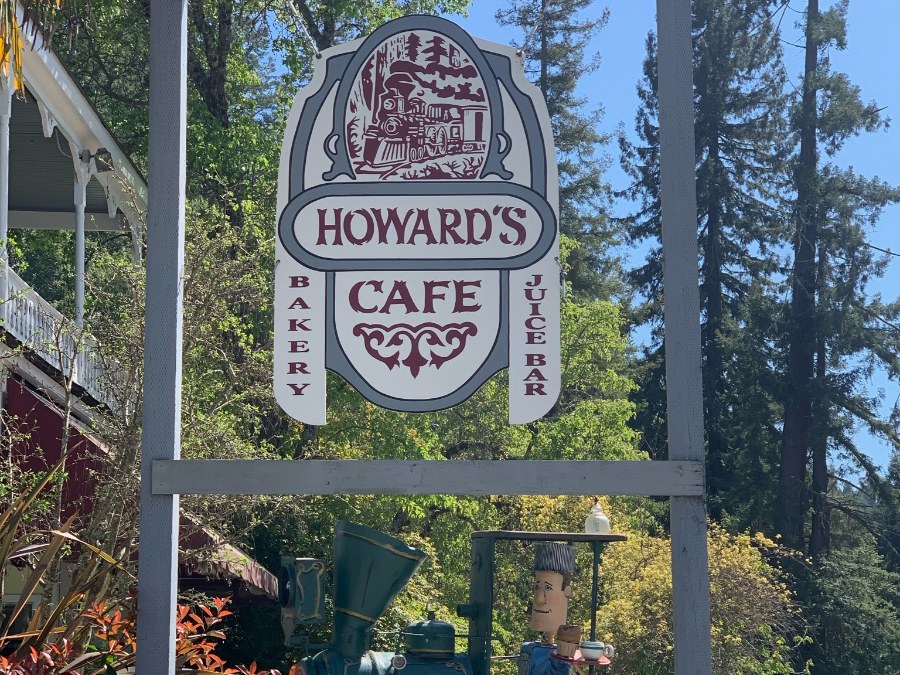 Sign of Howard's Cafe with a train illustration and train model beneath nearby trees. Link to Where to Eat on page.