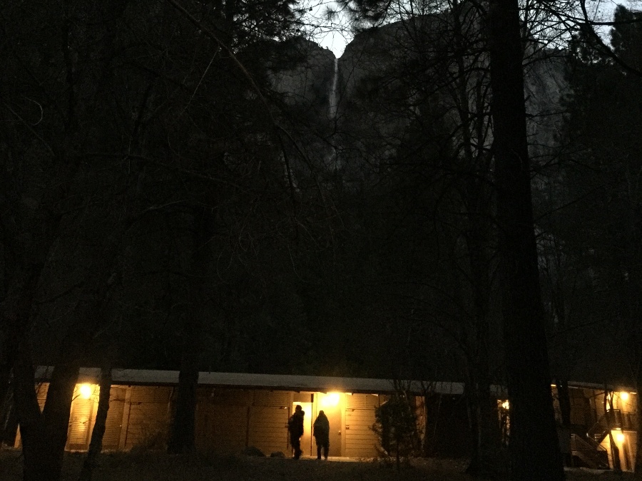 Dimly lit evening view of two silhouettes of people in front of Yosemite Valley Lodge with Yosemite Falls in background